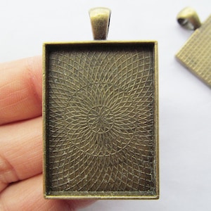 Könnte beinhalten: Ein quadratischer Anhänger aus bronzefarbenem Metall mit einem dekorativen, kreisförmigen Muster. Der Anhänger hat eine Öse zum Aufhängen.
