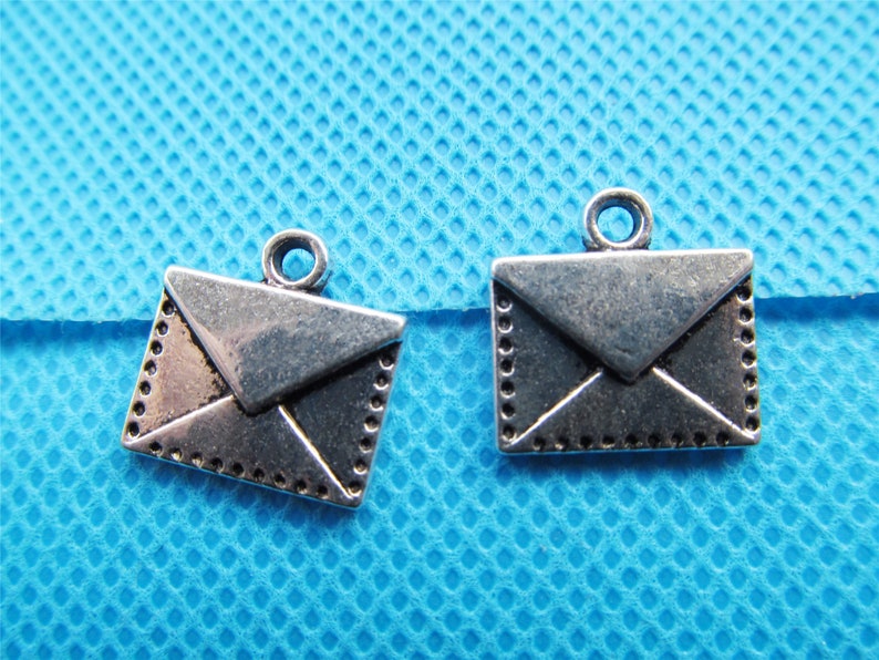 May include: Two silver-toned envelope charms with a decorative border. The charms have a loop at the top for attaching to a necklace or bracelet.