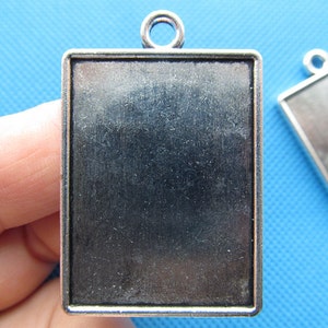 Puede incluir: Colgante de metal plateado con un marco rectangular y una superficie en blanco. El colgante tiene un bucle en la parte superior para sujetar una cadena o un cordón.