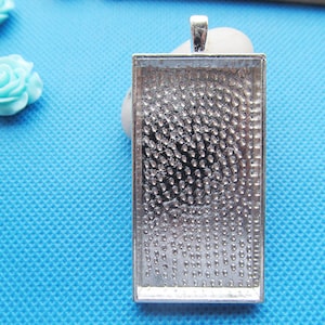 May include: Silver-toned pendant tray with a rectangular shape and a textured surface. The tray is designed to hold a small image or other decorative element.