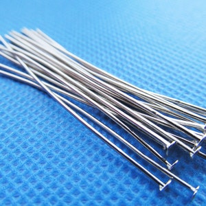 May include: A group of silver head pins with flat heads. The pins are arranged in a pile on a blue surface.