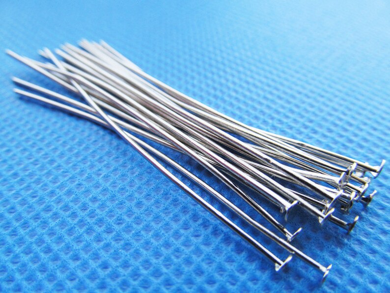 May include: A group of silver head pins with flat heads. The pins are arranged in a pile on a blue surface.