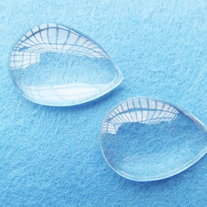 Op de afbeelding: Twee heldere glazen cabochons in druppelvorm op een lichtblauwe achtergrond. De cabochons zijn glad en hebben een licht afgeronde rand.