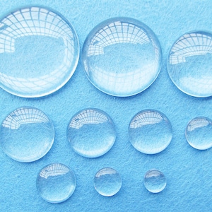 Peut inclure: Assortiment de cabochons en verre transparent de diff&eacute;rentes tailles, dispos&eacute;s sur une surface bleue. Les cabochons sont ronds et ont une finition lisse et polie.
