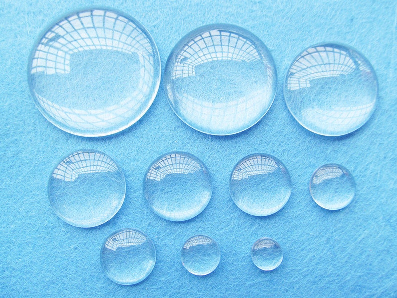 Peut inclure: Assortiment de cabochons en verre transparent de diff&eacute;rentes tailles, dispos&eacute;s sur une surface bleue. Les cabochons sont ronds et ont une finition lisse et polie.