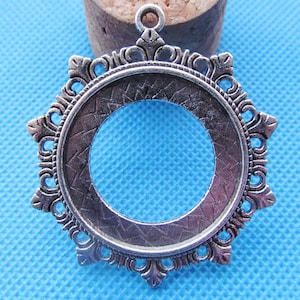 May include: Silver-toned metal pendant with a circular cutout and an ornate, decorative border. The pendant has a loop at the top for hanging.