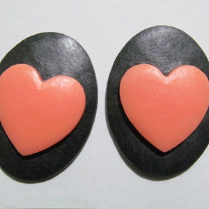 May include: A pair of black oval-shaped earrings with a raised pink heart design.