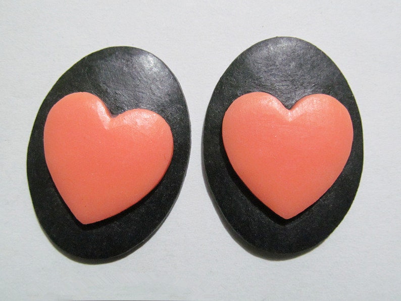 May include: A pair of black oval-shaped earrings with a raised pink heart design.