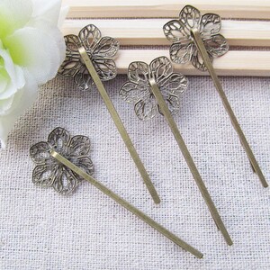 May include: Four bronze hair pins with decorative flower-shaped tops. The pins are arranged on a light brown fabric surface.
