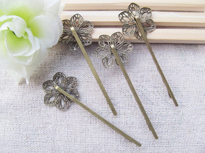 May include: Four bronze hair pins with decorative flower-shaped tops. The pins are arranged on a light brown fabric surface.