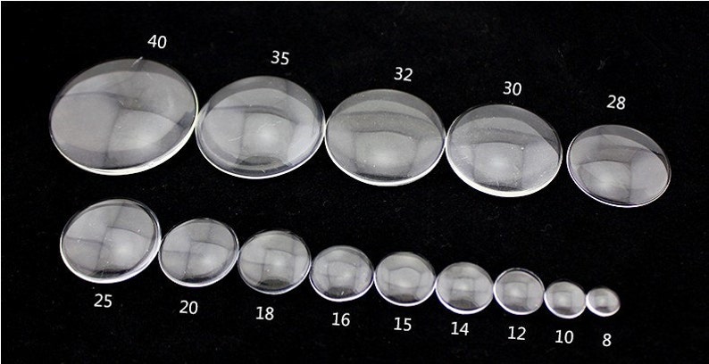 Peut inclure: Assortiment de cabochons en verre transparent de diff&eacute;rentes tailles, allant de 8 mm &agrave; 40 mm de diam&egrave;tre. Les cabochons sont dispos&eacute;s en deux rang&eacute;es, les plus grands en haut et les plus petits en bas.