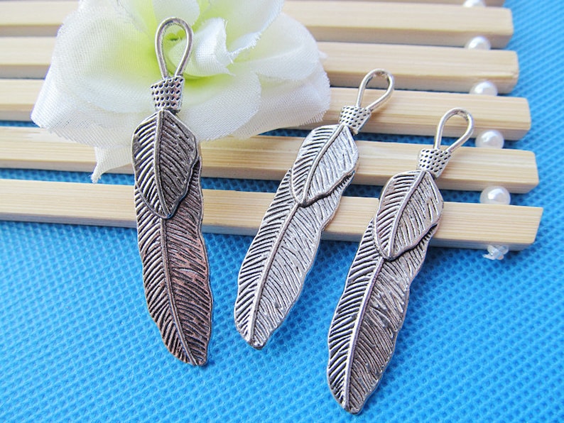May include: Three silver feather charms with a loop at the top for attaching to jewelry or other crafts. The feathers are detailed with a textured surface and have a slightly oxidized finish.