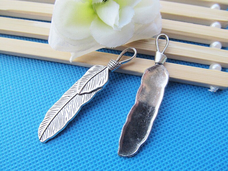 May include: Two silver-toned metal charms in the shape of feathers. One charm is a simple, flat feather, while the other is a more detailed, textured feather.