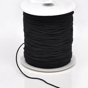 Könnte beinhalten: Eine Spule mit schwarzem Gummiband, das um eine weiße Kunststoffspule gewickelt ist. Das Band ist dünn und flexibel und eignet sich ideal zum Basteln und für die Schmuckherstellung.