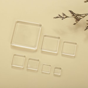 以下が含まれることがあります： さまざまなサイズの透明な正方形のガラスタイル。 タイルは、右上隅に乾燥した葉の小枝が付いた、明るいベージュの表面に配置されています。 ガラスタイルは透明で、光を反射します。