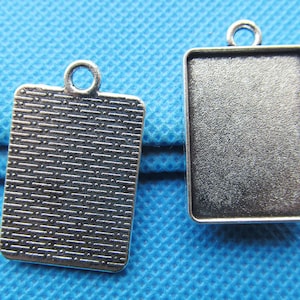 May include: Two silver-toned pendant trays with a rectangular shape. The tray on the left has a textured surface and a loop at the top for hanging. The tray on the right is smooth and has a loop at the top for hanging.