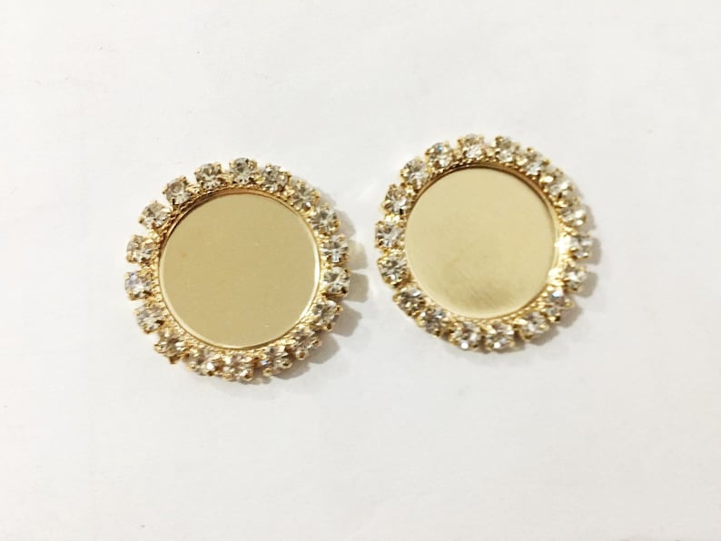 May include: Two gold-toned round metal frames with clear rhinestones around the edges. The frames are blank and can be used for crafting.