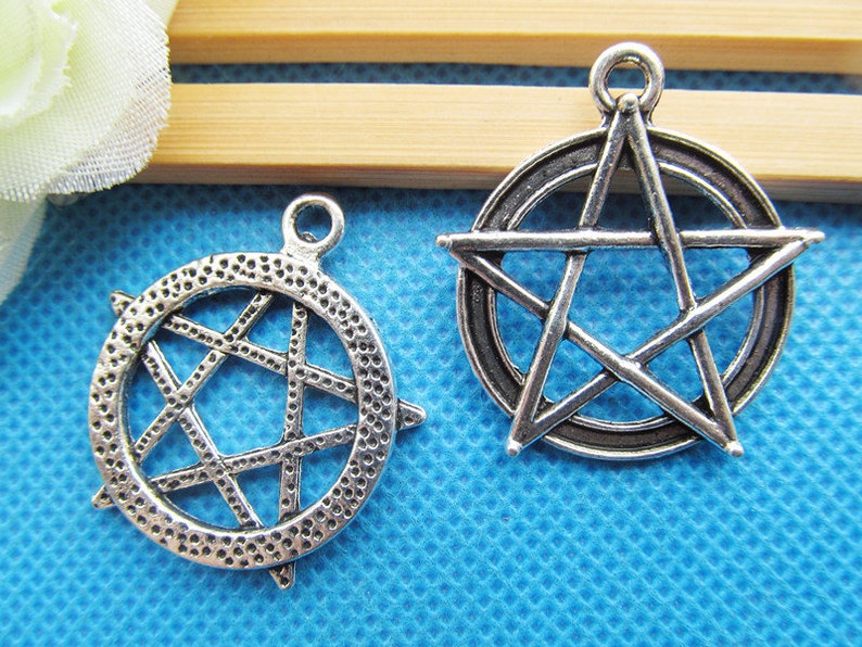 May include: Two silver-toned metal charms in the shape of a five-pointed star. The star on the left is enclosed in a circle with a textured surface. The star on the right is outlined with a circle.