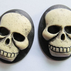 May include: Two white skull-shaped buttons with black outlines. The skulls have black eye sockets and a toothy grin.