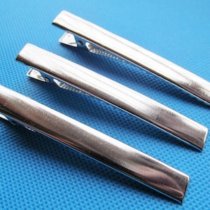 Puede incluir: Tres pinzas para el pelo de metal plateado, cada una con una superficie lisa ligeramente texturizada. Las pinzas están dispuestas en fila, con la pinza superior superponiéndose ligeramente a la pinza del medio.