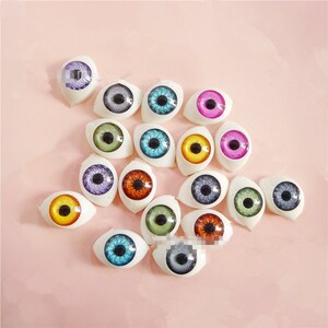 Op de afbeelding: Een verzameling poppenogen in verschillende kleuren, waaronder blauw, groen, geel, paars en oranje. Elk oog heeft een gedetailleerde iris en pupil, geplaatst in een witte, ovale basis. De ogen zijn gerangschikt op een roze oppervlak.