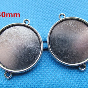 Può includere: Due basi per ciondoli in metallo color argento, ciascuna con una superficie rotonda e liscia di 30 mm di diametro. Le basi hanno un anello nella parte superiore per attaccare una catena o un cordino.