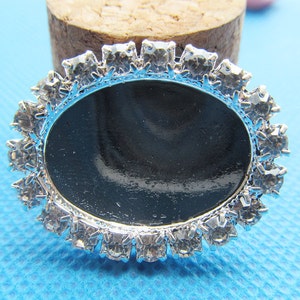 May include: Silver oval-shaped pendant with a black background and a clear rhinestone border.