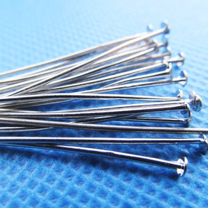 May include: A close-up of a pile of silver-toned head pins. The pins are arranged in a fan shape on a blue textured surface.