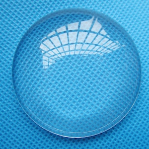 May include: A clear glass magnifying lens, also known as a reading glass, on a blue textured surface.