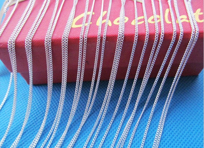 May include: A close-up image of a large quantity of silver chains, arranged in rows, on a red box with the word "Chocalat" printed on it. The chains are lying on a blue surface.