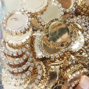 May include: A close-up of a pile of gold-toned metal embellishments with clear rhinestones. The embellishments are round and have a flat surface in the center.