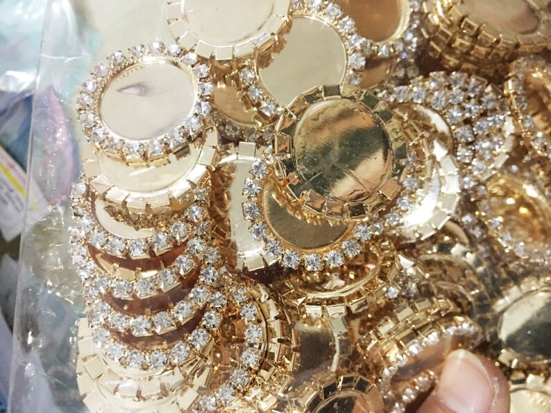 May include: A close-up of a pile of gold-toned metal embellishments with clear rhinestones. The embellishments are round and have a flat surface in the center.