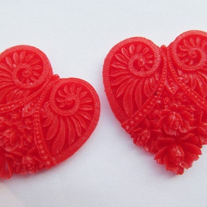 Può includere: Due bottoni rossi a forma di cuore con intricati disegni floreali e a spirale. I bottoni sono realizzati in un materiale lucido simile alla plastica.