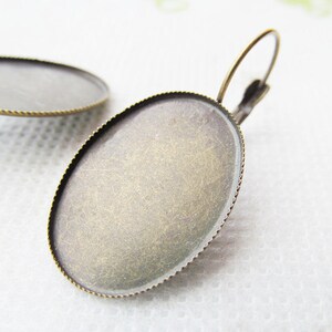 May include: A pair of oval-shaped earring blanks with a brass finish. The blanks have a raised edge and a smooth surface. The earrings are ready to be decorated with your favorite embellishments.