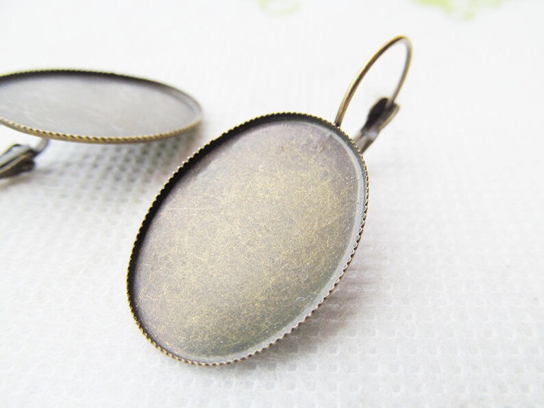 May include: A pair of oval-shaped earring blanks with a brass finish. The blanks have a raised edge and a smooth surface. The earrings are ready to be decorated with your favorite embellishments.
