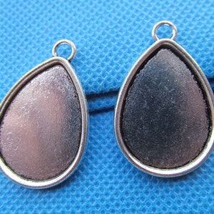 Könnte beinhalten: Zwei silberne Anhänger in Tropfenform mit einer glatten, leicht strukturierten Oberfläche. Die Anhänger sind so konzipiert, dass sie einen Cabochon-Stein oder einen anderen Schmuckstein aufnehmen können.