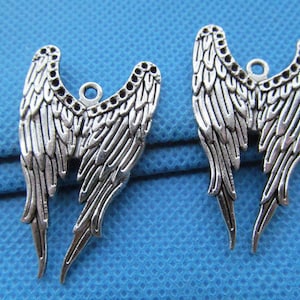 May include: Two silver angel wing charms with a detailed design. The wings are connected at the top and have a hole for attaching to a necklace or bracelet.