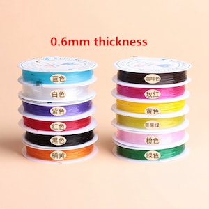 May include: A stack of spools of elastic string in various colors, including blue, white, purple, red, black, orange, brown, pink, yellow, and green. The spools are labeled with the color names in Chinese characters. The text "0.6mm thickness" is printed above the spools.