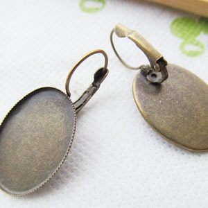 May include: A pair of antique gold-toned earring blanks with a round, slightly oval-shaped bezel. The bezels are smooth and have a slightly textured surface. The earring hooks are made of metal and have a spring-loaded closure.