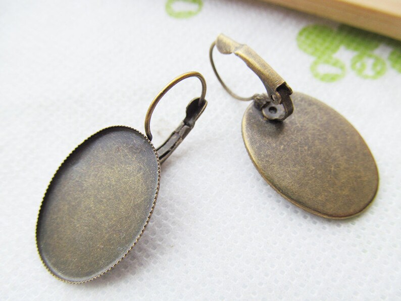 May include: A pair of antique gold-toned earring blanks with a round, slightly oval-shaped bezel. The bezels are smooth and have a slightly textured surface. The earring hooks are made of metal and have a spring-loaded closure.