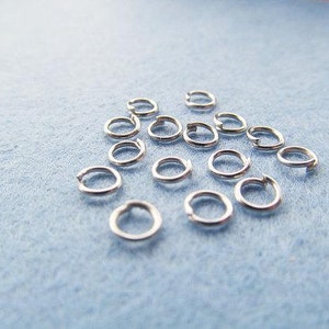 May include: A collection of silver jump rings, a type of jewelry finding, scattered on a light blue surface.
