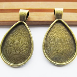 Op de afbeelding: Twee bronzen hanger-schalen in een traanvorm met een getextureerd oppervlak. De schalen zijn ontworpen om een klein stukje glas of ander materiaal te bevatten.