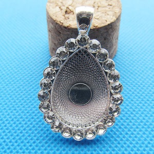 Könnte beinhalten: Silberfarbener Anhänger in Tropfenform, umgeben von klaren, runden Kristallen. Der Anhänger hat ein strukturiertes Innenmuster und eine kleine Schlaufe für eine Kette. Der Hintergrund ist hellblau.