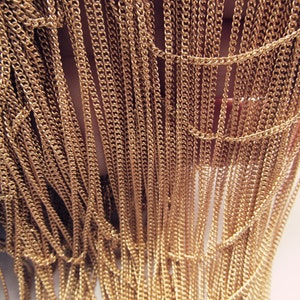 May include: A close-up of a gold chain fringe. The fringe is made of many thin chains that are all connected together. The chains are hanging down in a curtain-like fashion.
