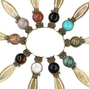Puede incluir: Conjunto de diez marcapáginas con forma de búho con acentos de piedras preciosas de diferentes colores. Cada marcapáginas presenta un diseño de búho detallado con una piedra preciosa redonda en el centro. Las piedras preciosas incluyen rojo, negro, turquesa, marrón, ojo de tigre, blanco, azul, verde y dos tonos diferentes de marrón.
