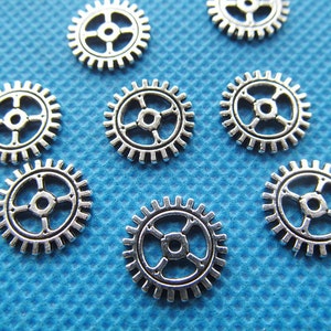 May include: A collection of eight silver-toned metal gears of varying sizes. The gears are arranged on a blue textured surface.