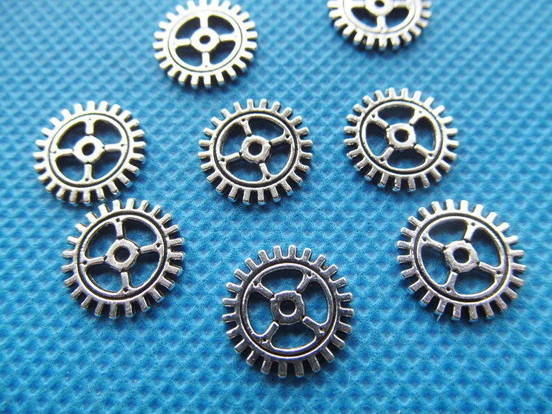 May include: A collection of eight silver-toned metal gears of varying sizes. The gears are arranged on a blue textured surface.