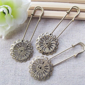 May include: Three antique gold safety pins with decorative, ornate, circular, filigree designs. The pins are on a light brown fabric background.