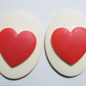 May include: Two white oval-shaped earrings with a red heart design in the center of each earring.