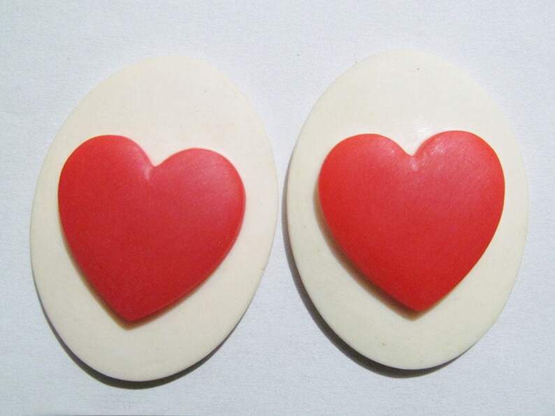 May include: Two white oval-shaped earrings with a red heart design in the center of each earring.